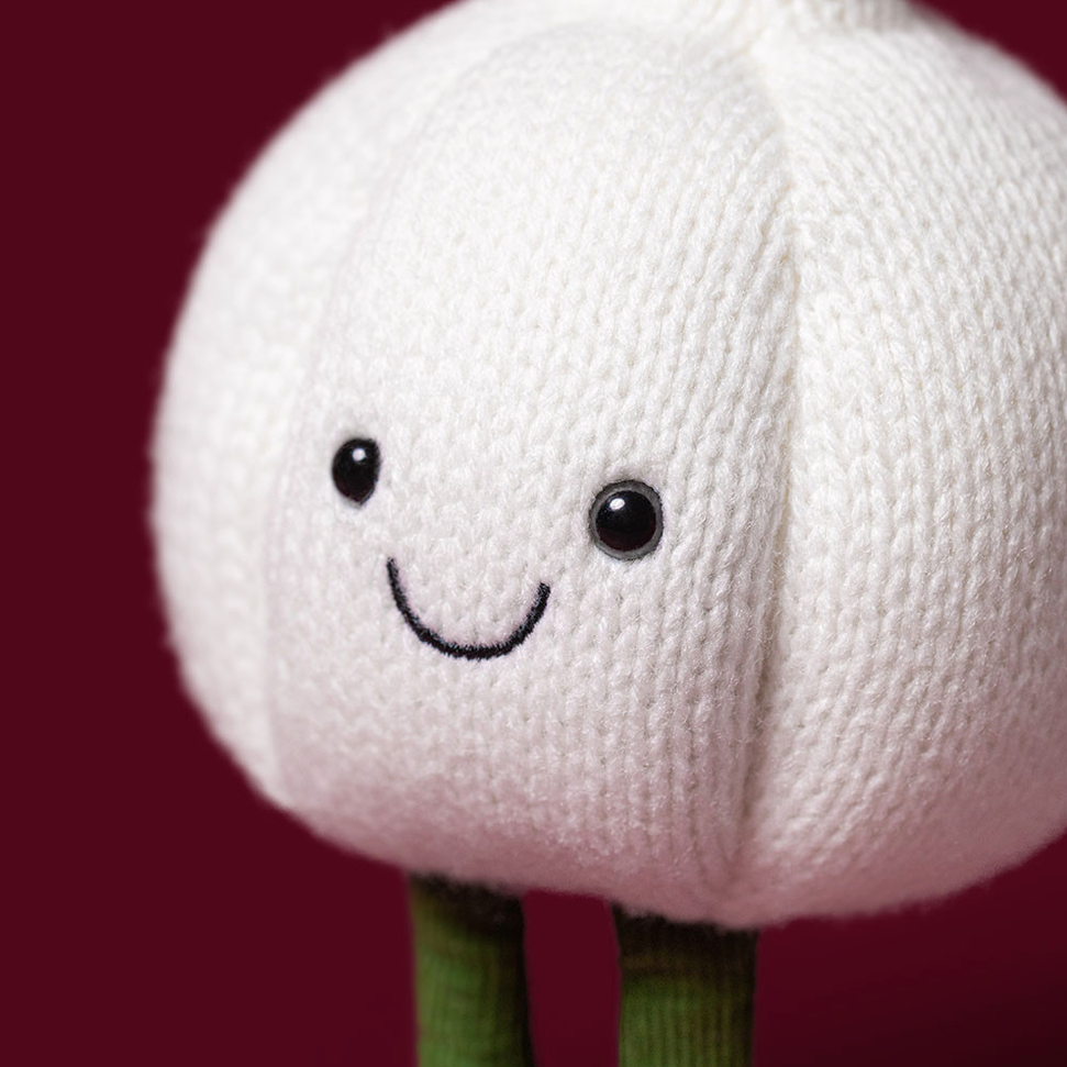 A close-up of a knitted garlic character toy with a smiling face, black bead eyes, and green stems against a dark burgundy background.