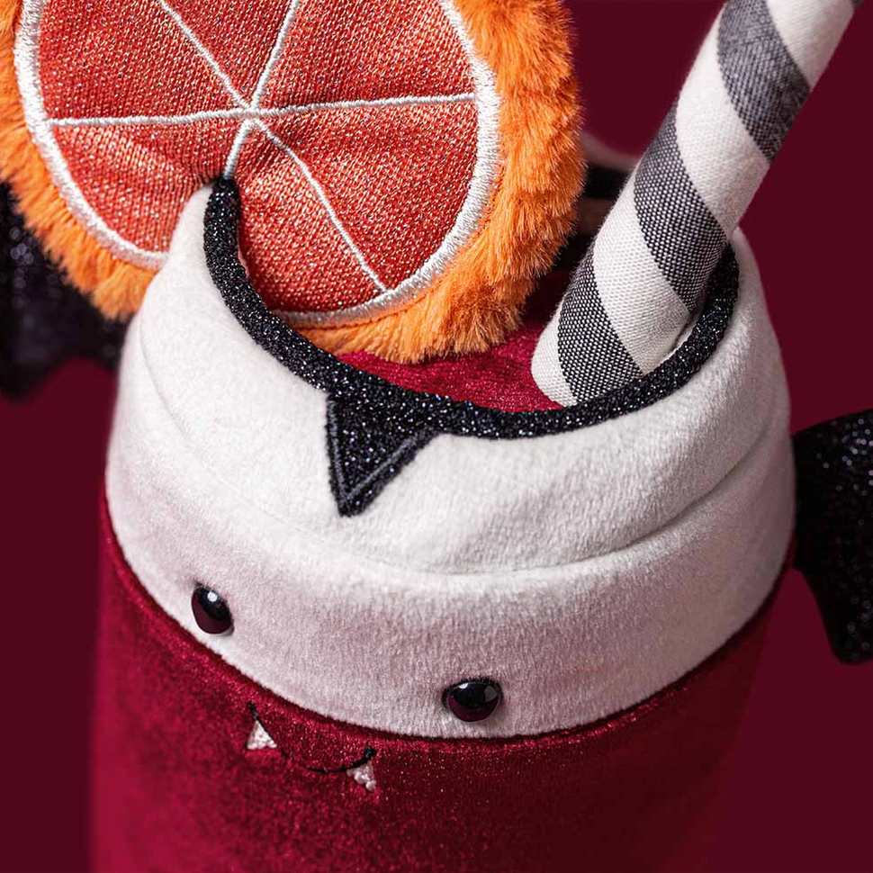 A close-up of a plush drink cup accessory with a white top, red glittery lower section, black bat-wing details, a striped straw, and an orange slice decoration on a dark red background.