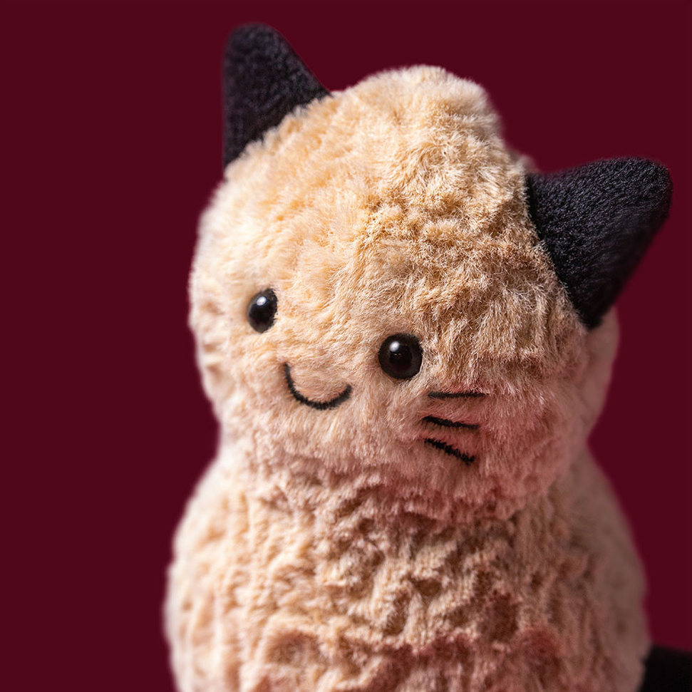 A close-up of a plush cat toy with cream-colored textured fur, black ears, and a stitched smiling face, set against a solid dark burgundy background.