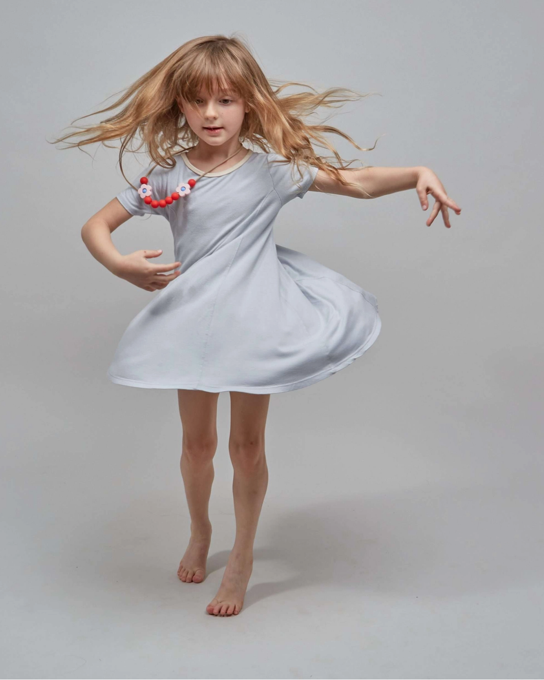 A young girl with long blonde hair twirling in a light gray short-sleeve dress, wearing a red and white beaded necklace, against a solid light gray studio background.