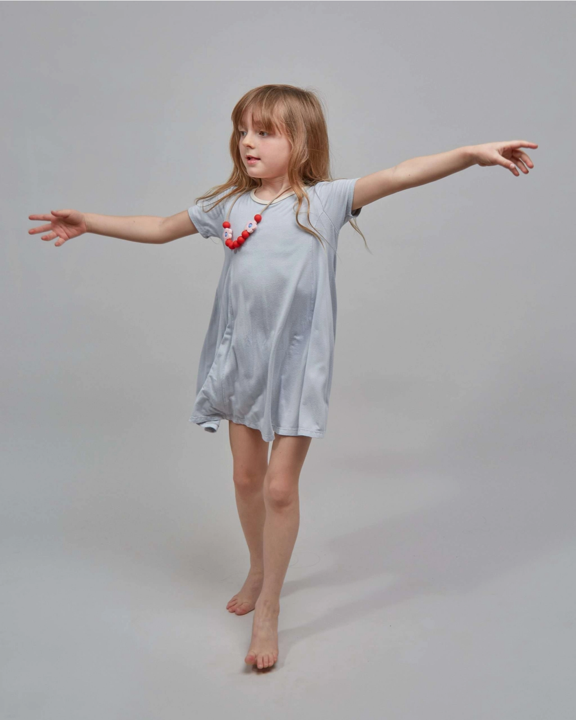 A young girl with long light brown hair poses in a light blue-gray short-sleeve tunic dress, accessorized with a red and white beaded necklace, against a solid light gray studio background.