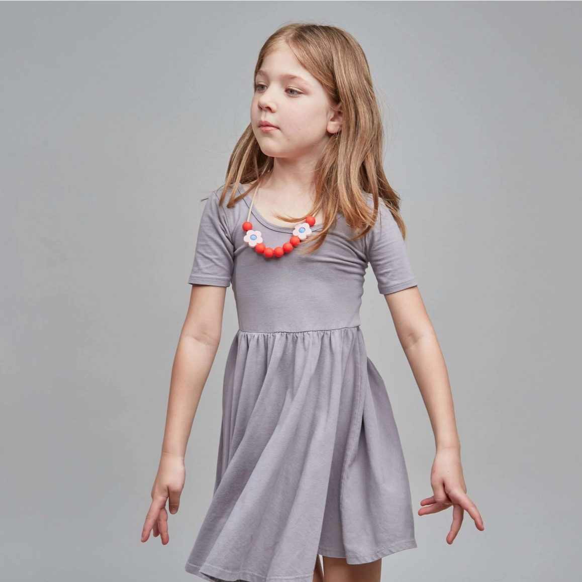 A young girl with blonde hair wearing a short-sleeved gray dress and a red and white beaded necklace, standing against a solid gray studio background.
