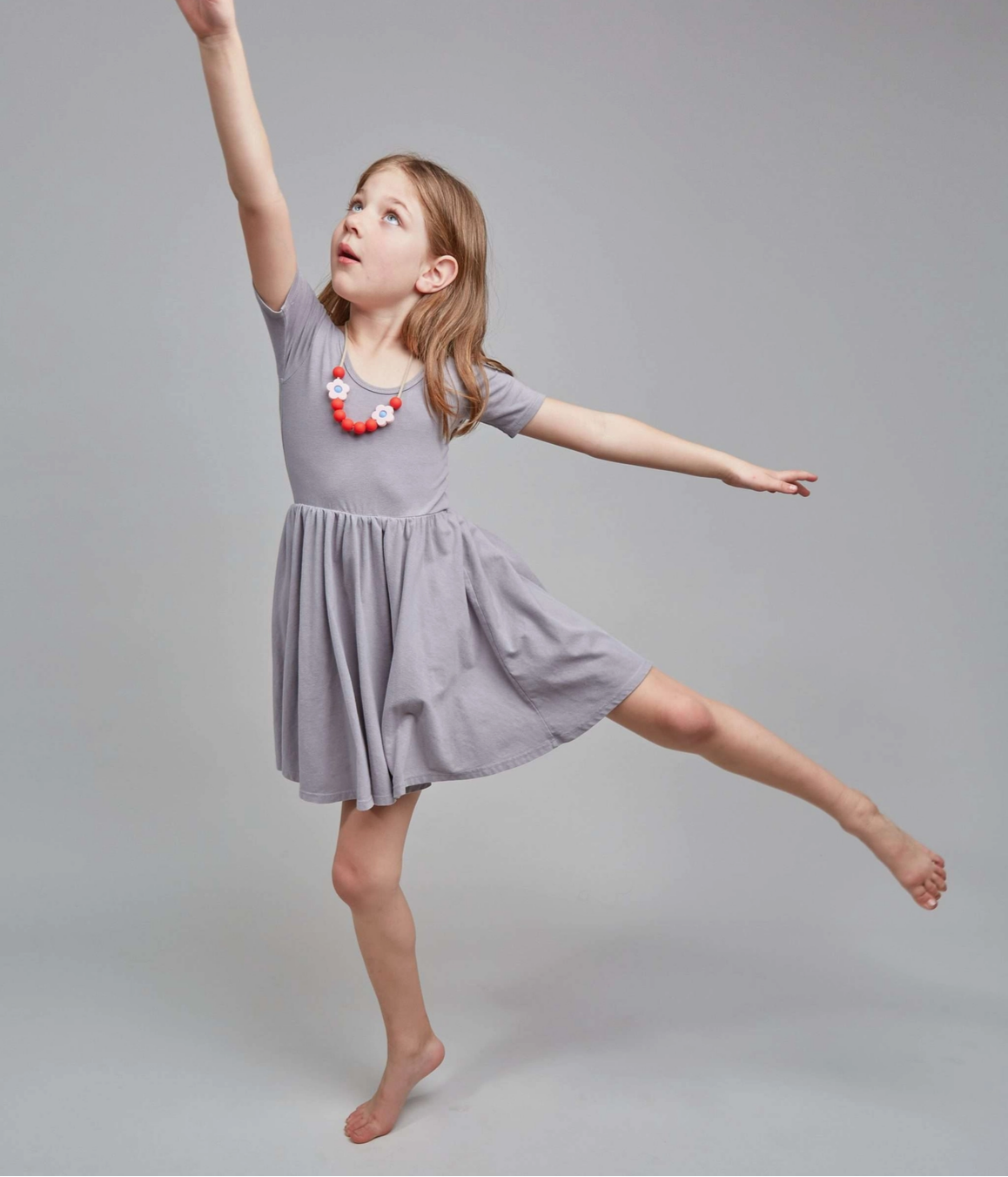 A young girl in a light gray short-sleeve dress and a colorful beaded necklace poses dynamically with one arm raised and the other extended, set against a solid gray studio background.