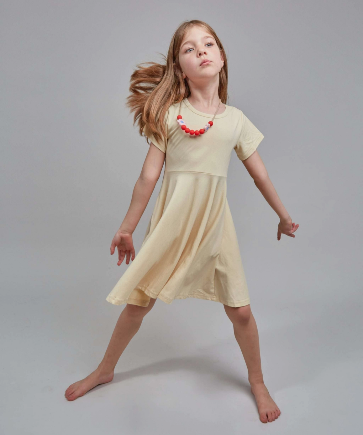 A young girl with long light brown hair poses in a light beige short-sleeve dress, accessorized with a red beaded necklace, against a solid gray studio background.