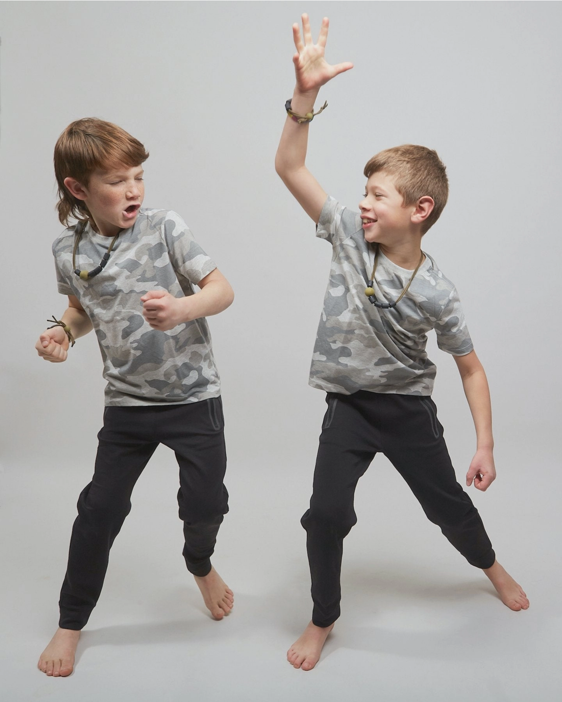 Two young boys modeling matching gray camouflage t-shirts and black jogger pants, posing dynamically against a light gray studio background.