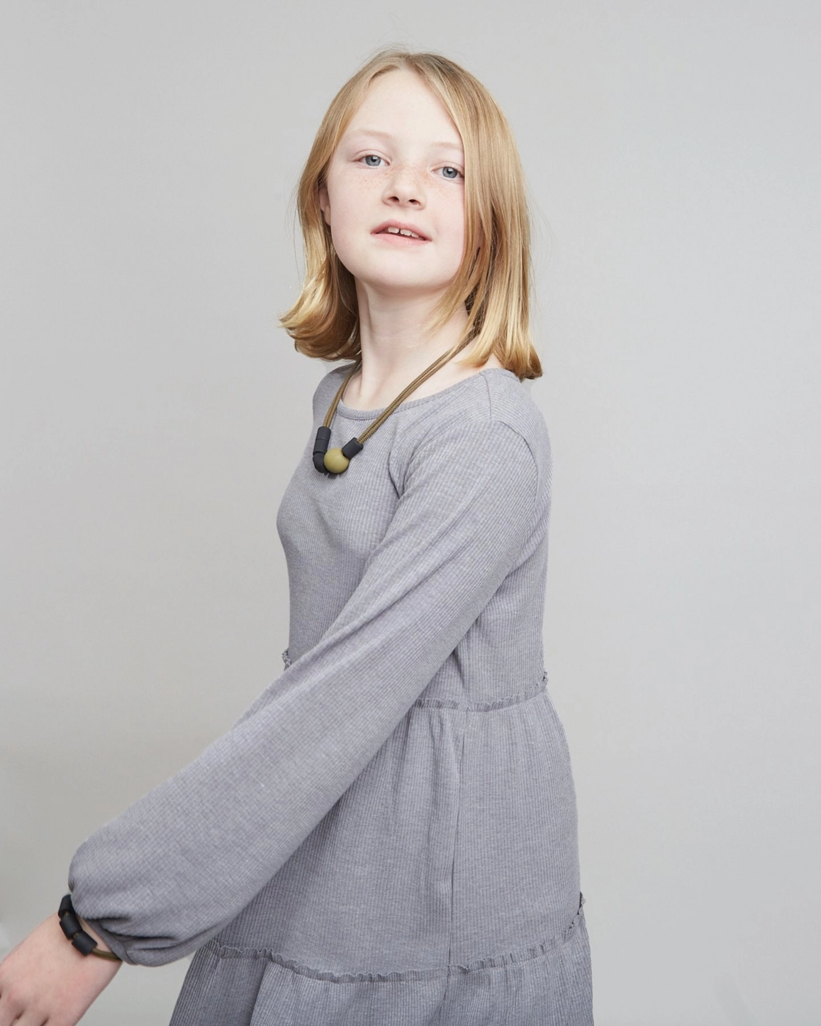 A young girl with blonde hair wearing a gray long-sleeve dress and a brown beaded necklace, photographed against a solid light gray background.