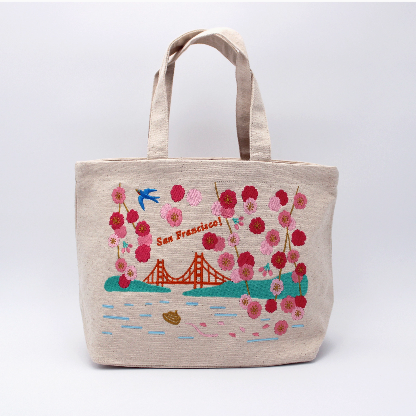 A beige canvas tote bag featuring a colorful embroidered design of the Golden Gate Bridge, pink and red cherry blossoms, a blue bird, and the text 'San Francisco!' on a light gray studio background.