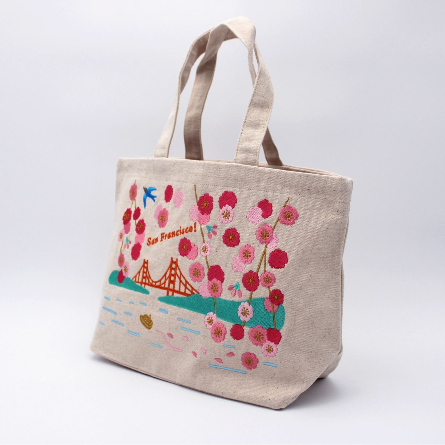 A beige canvas tote bag featuring a colorful embroidered San Francisco design with cherry blossoms, the Golden Gate Bridge, and the text 'San Francisco!' on a light gray studio background.