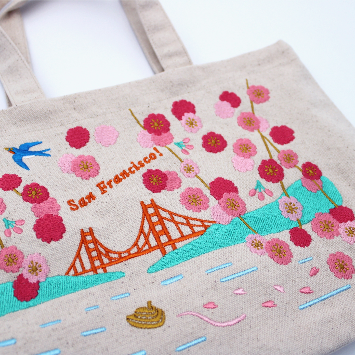 A beige canvas tote bag with embroidered San Francisco-themed artwork, including the Golden Gate Bridge, pink cherry blossoms, a blue bird, and the text 'San Francisco!'.
