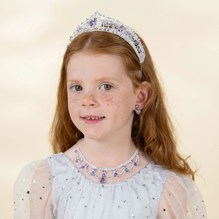 A young girl with freckles and red hair is shown from the chest up, wearing a silver tiara with blue and purple crystals, a matching crystal necklace, and a light blue dress against a cream background.