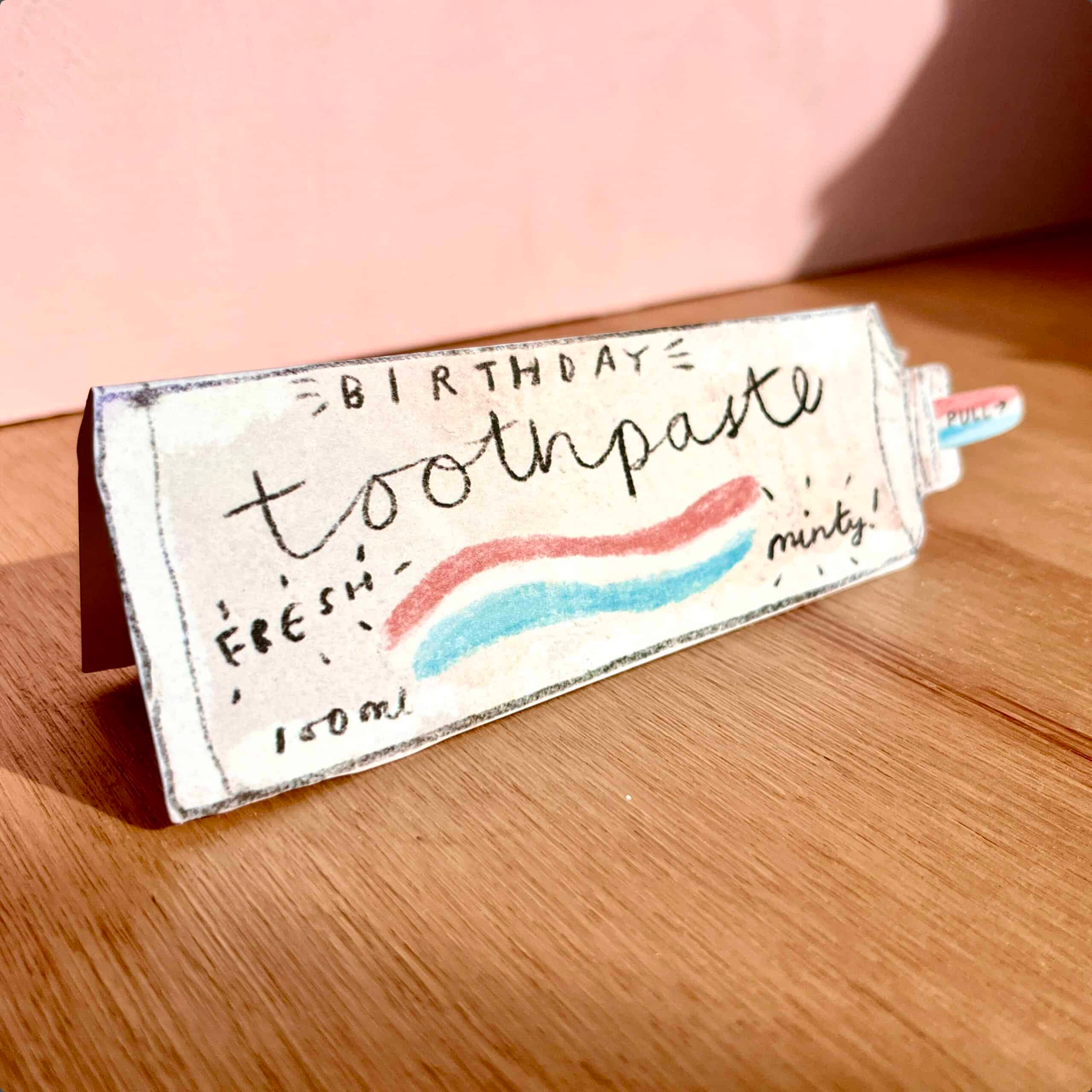 A novelty toothpaste tube-shaped card  with hand-lettered text reading 'BIRTHDAY toothpaste' and a pink and blue wave design, photographed on a wooden tabletop against a soft pink background.