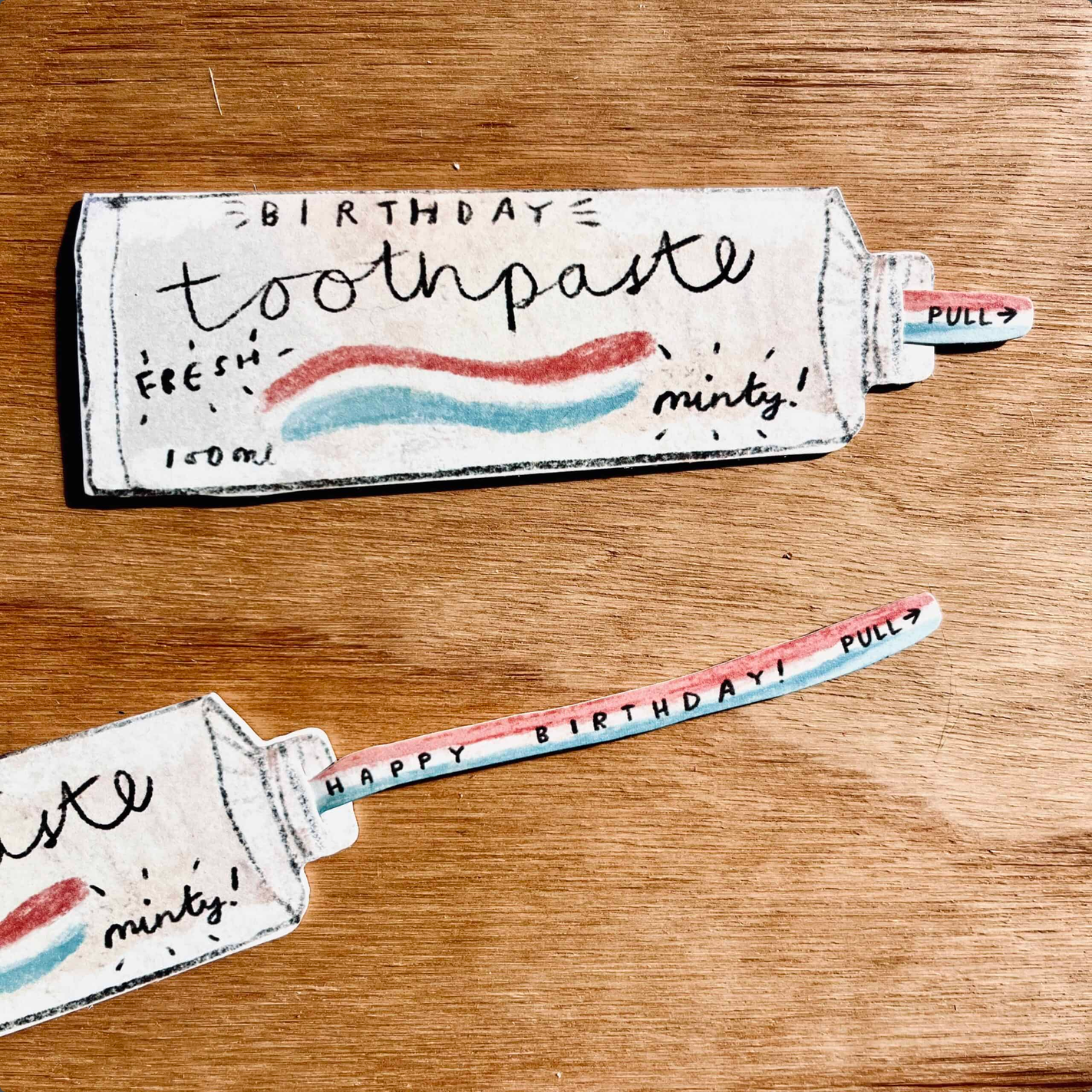 A novelty toothpaste-shaped paper pull with a printed 'BIRTHDAY toothpaste' design, shown on a wooden tabletop. The pull has a white base with hand-drawn red and blue toothpaste stripes and black text, plus a pull tab labeled 'PULL'.