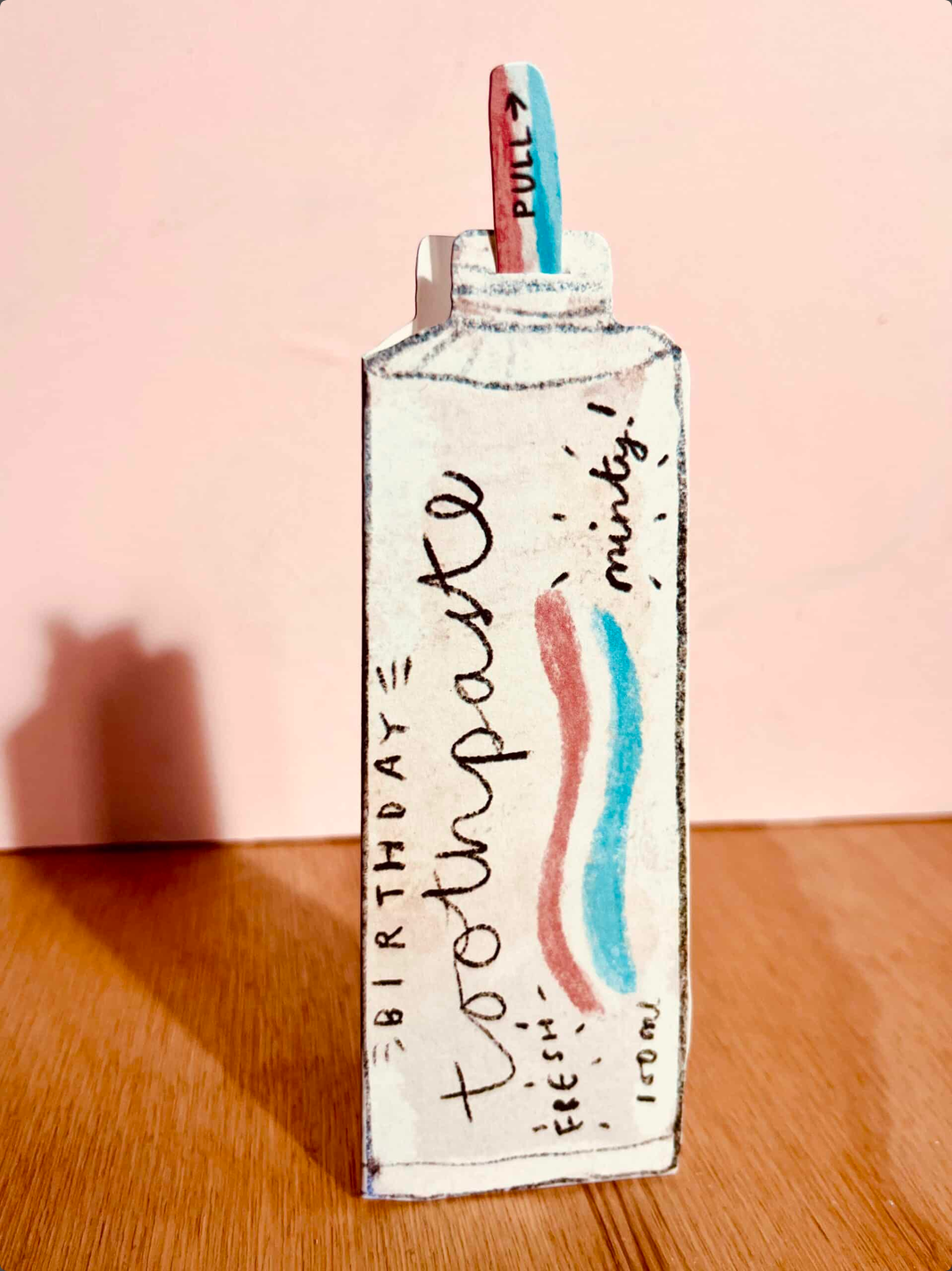 A handmade cardboard toothpaste bottle card with 'toothpaste' written on the front, decorated with red and blue toothpaste swirls and a pull tab, standing on a wooden surface against a pink background.