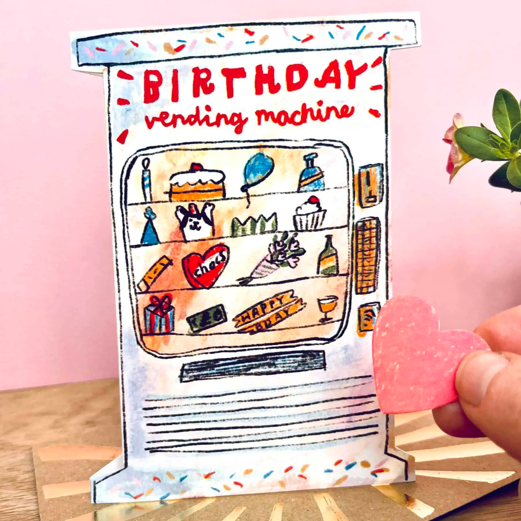 A handmade paper  card shaped like a retro vending machine with 'BIRTHDAY vending machine' written on it, displayed on a gold-patterned card with a pink background. A hand is holding a pink heart-shaped candy near the machine.