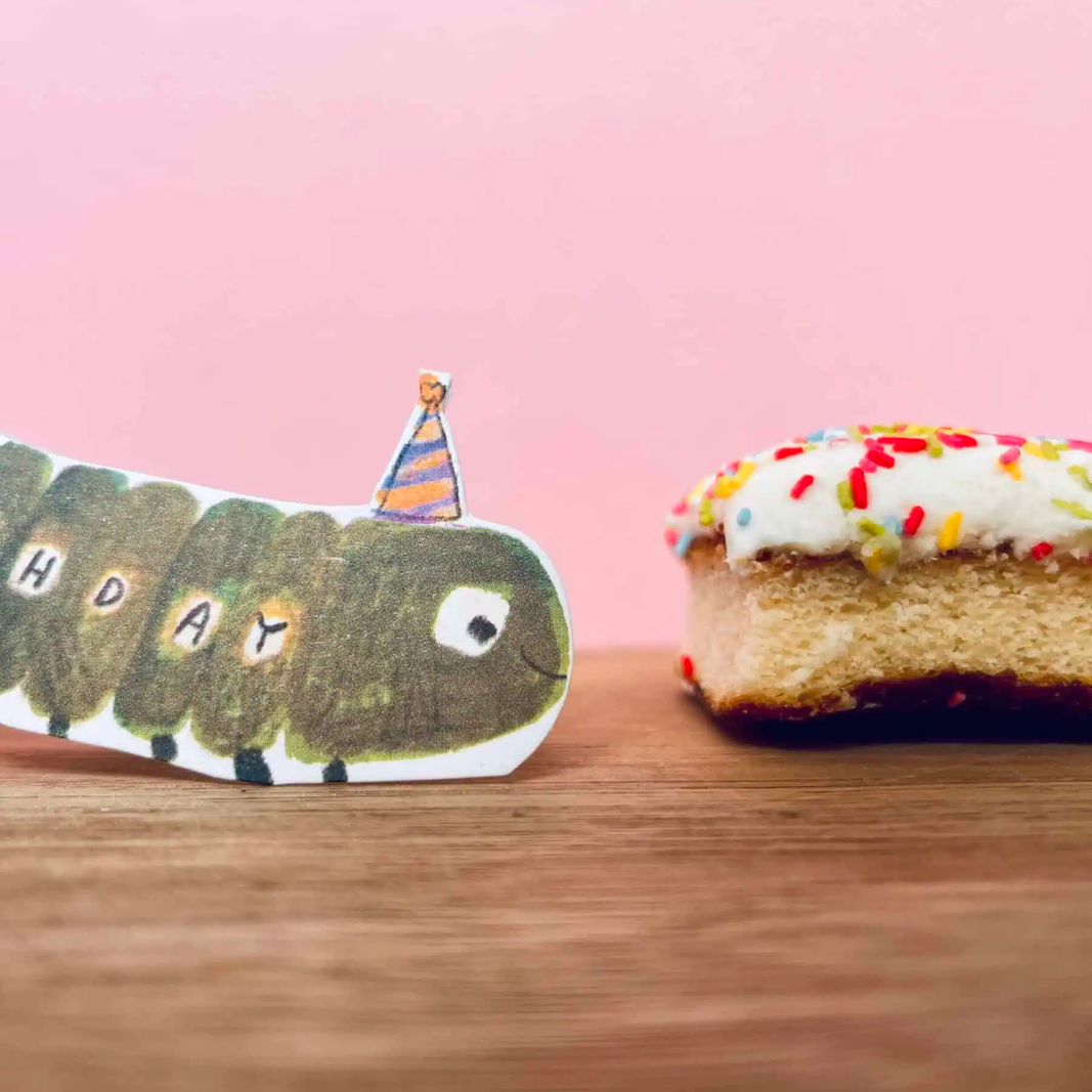 A handmade paper caterpillar card with the word 'HAPPY' spelled across its body and a small striped party hat, placed on a wooden surface next to a slice of cake with white frosting and colorful sprinkles against a pink background.