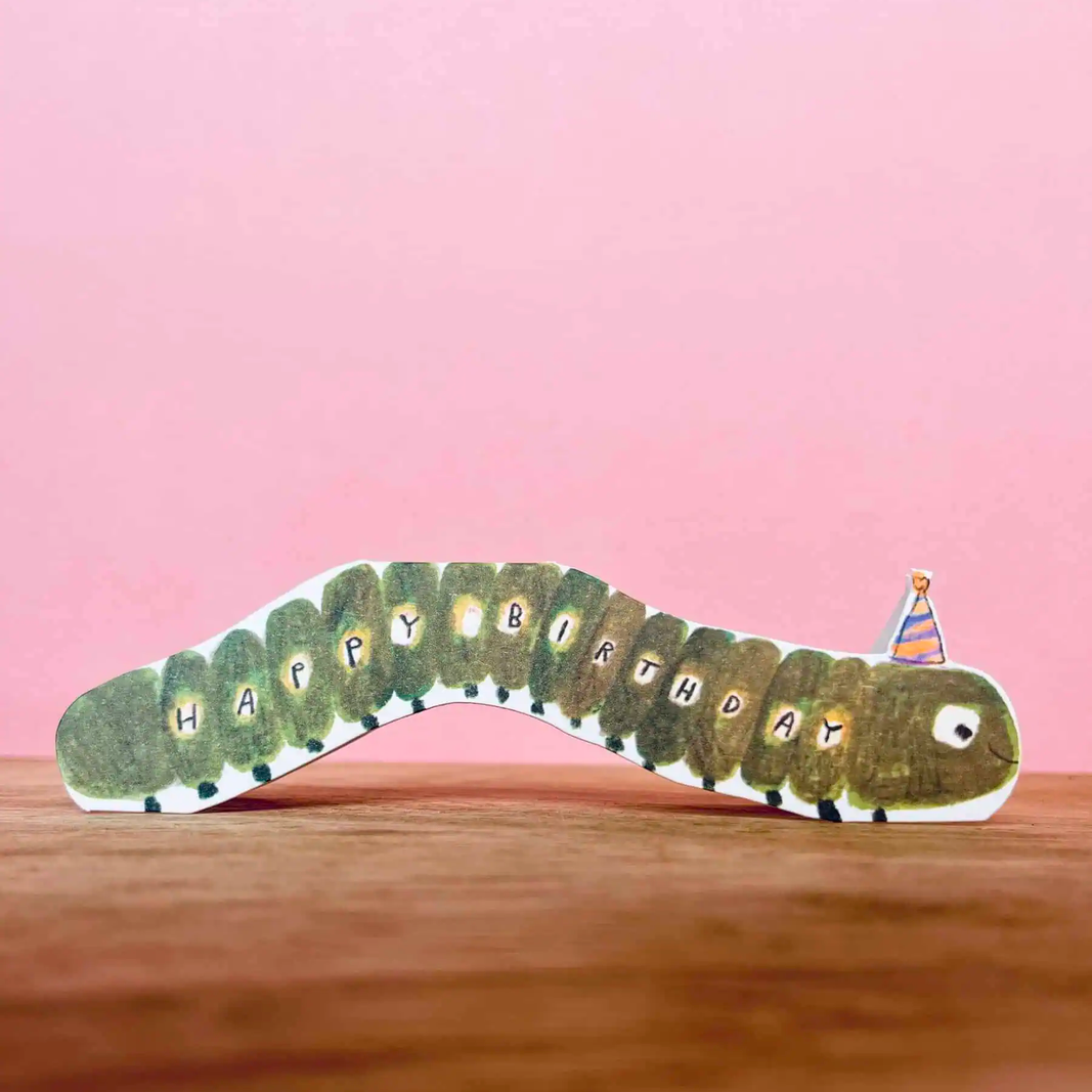 A handmade paper caterpillar birthday card or decoration with 'HAPPY BIRTHDAY' spelled out on its back and a small party hat, resting on a wooden surface against a pink background.