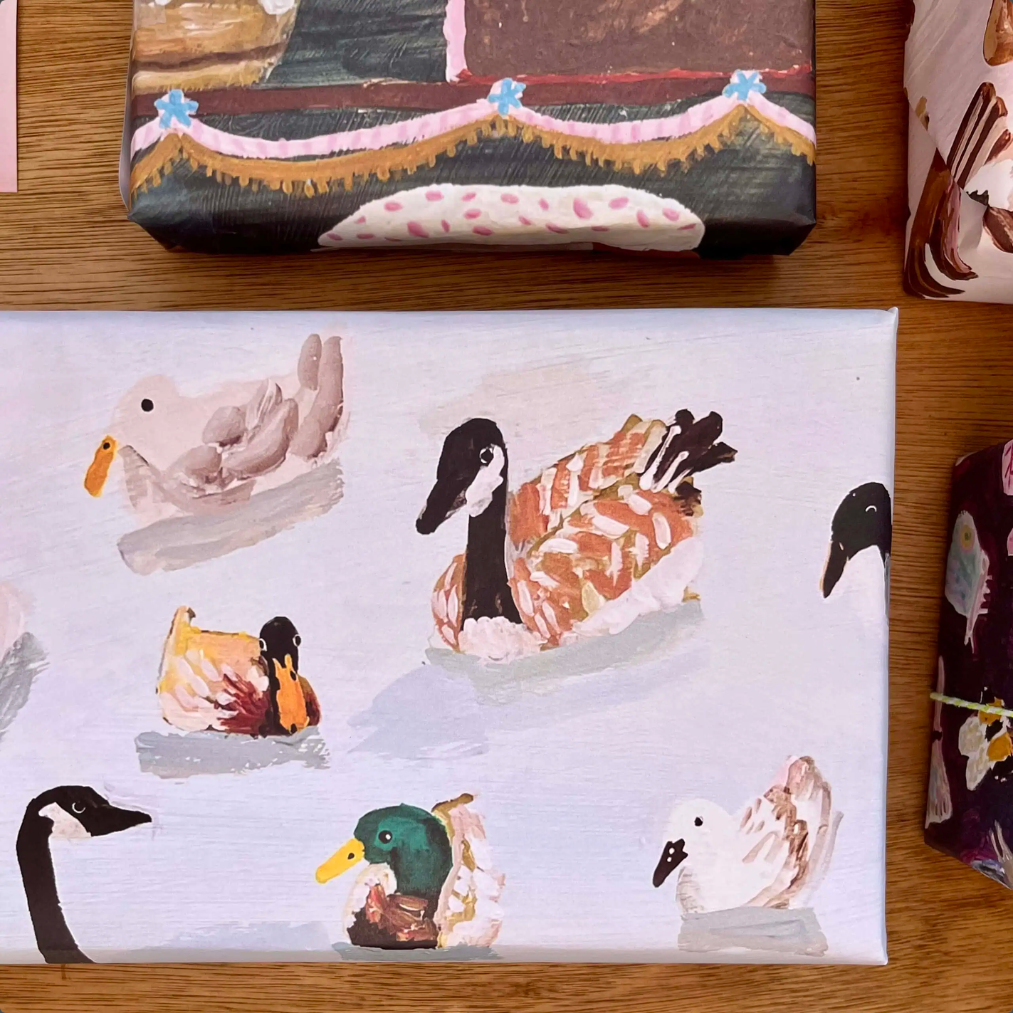 A rectangular canvas print featuring a painted pattern of various ducks and geese swimming on water, displayed on a wooden surface next to other wrapped gift boxes.