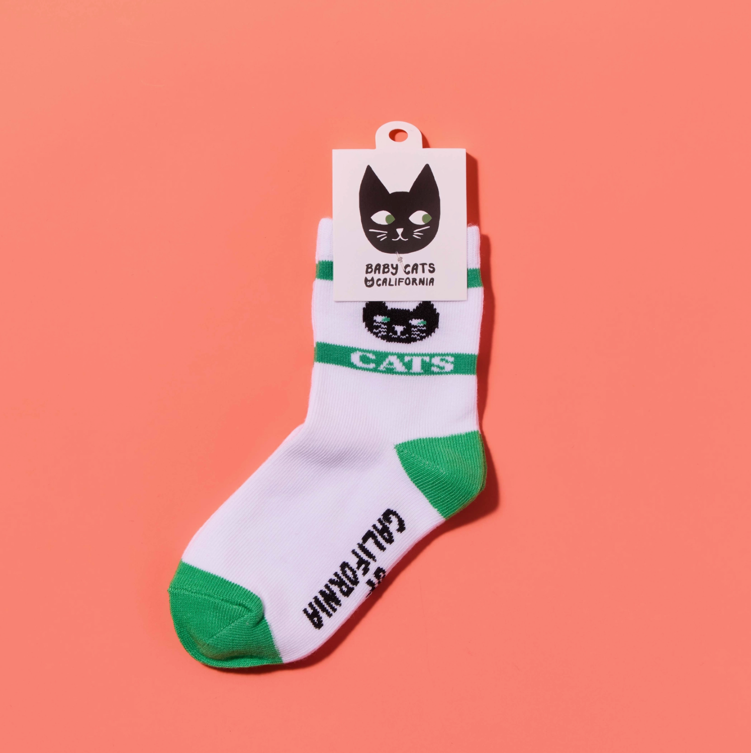 A pair of white crew socks with green toes and heels, featuring a green stripe with 'CATS' and a black cat face logo, along with 'BABY CATS CALIFORNIA' printed on the attached paper tag. The socks are displayed against a solid coral pink background.