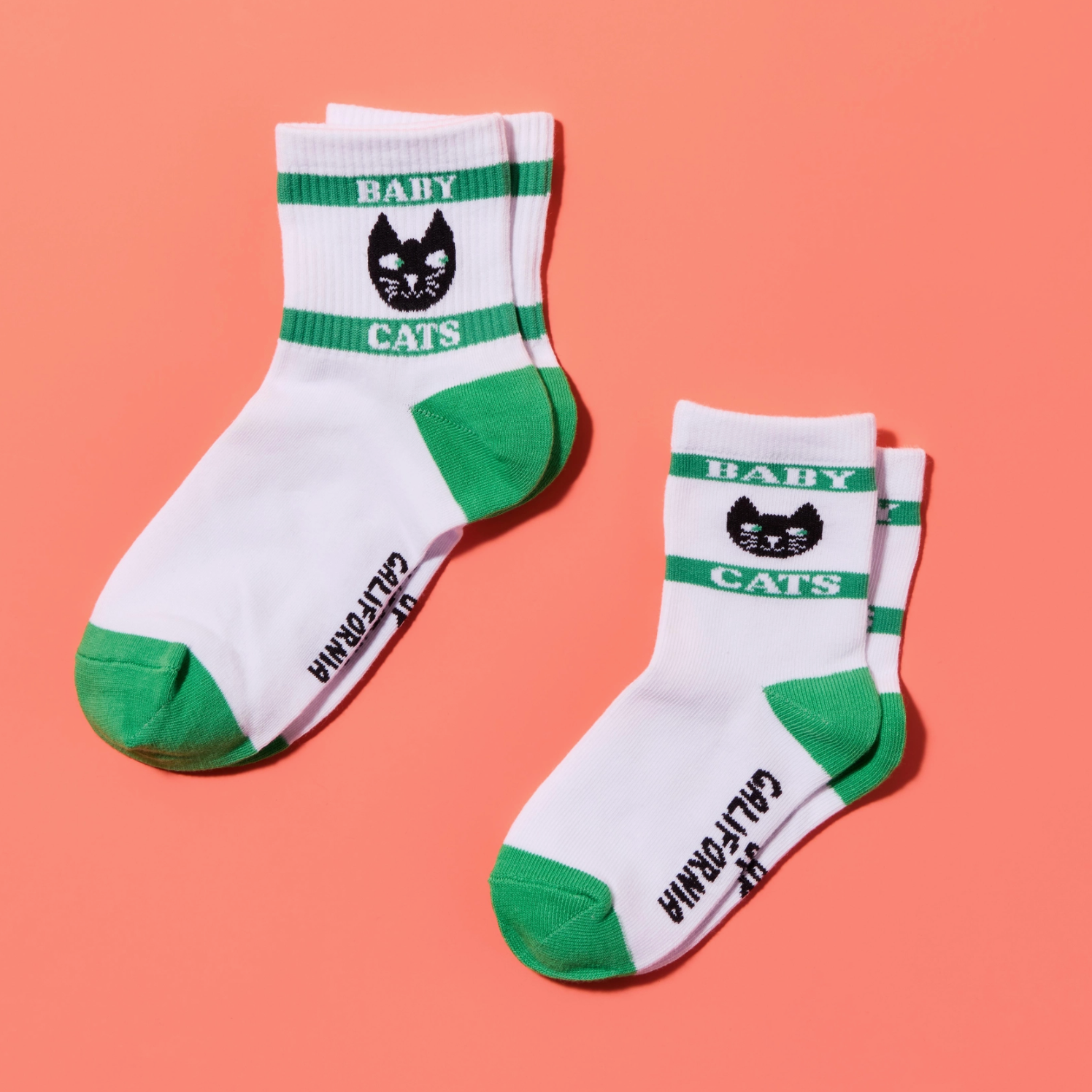Two white crew socks with green toe, heel, and cuff accents, featuring a black cat face logo, the words 'BABY CATS' on the cuff, and 'CALIFORNIA' printed vertically near the ankle. The socks are arranged on a solid coral pink background.