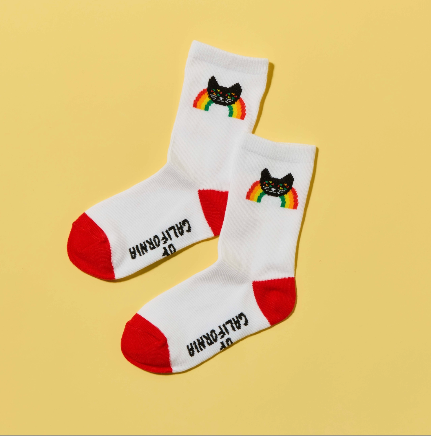 A pair of white crew socks with red toes and heels, featuring a black cat and rainbow graphic near the cuff, with 'CALIFORNIA' printed in black. The socks are photographed on a solid yellow background.