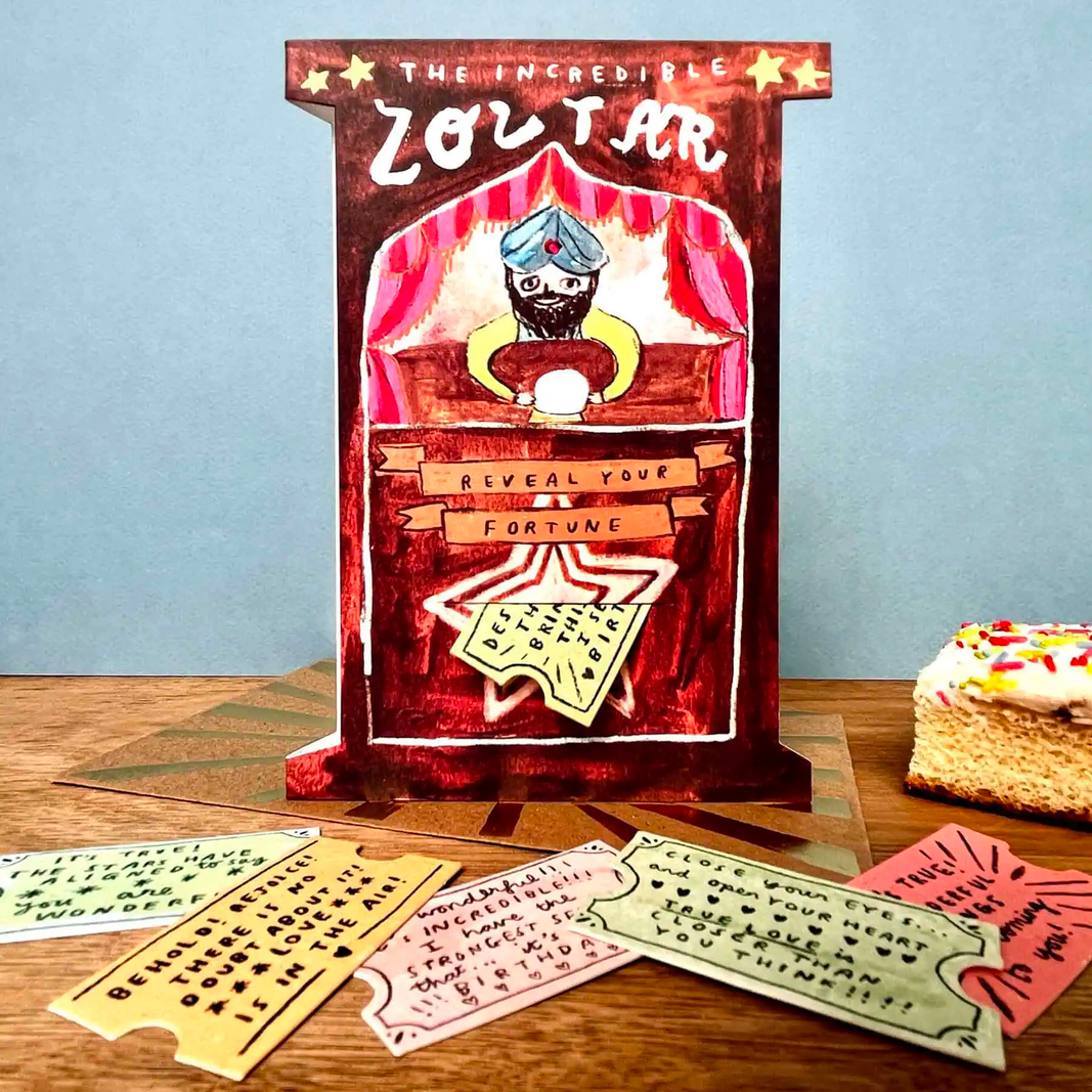 A hand-drawn style Zoltar fortune card dck displayed on a wooden table against a light blue background. The main card features a magician character with a blue turban and black beard behind a red podium.