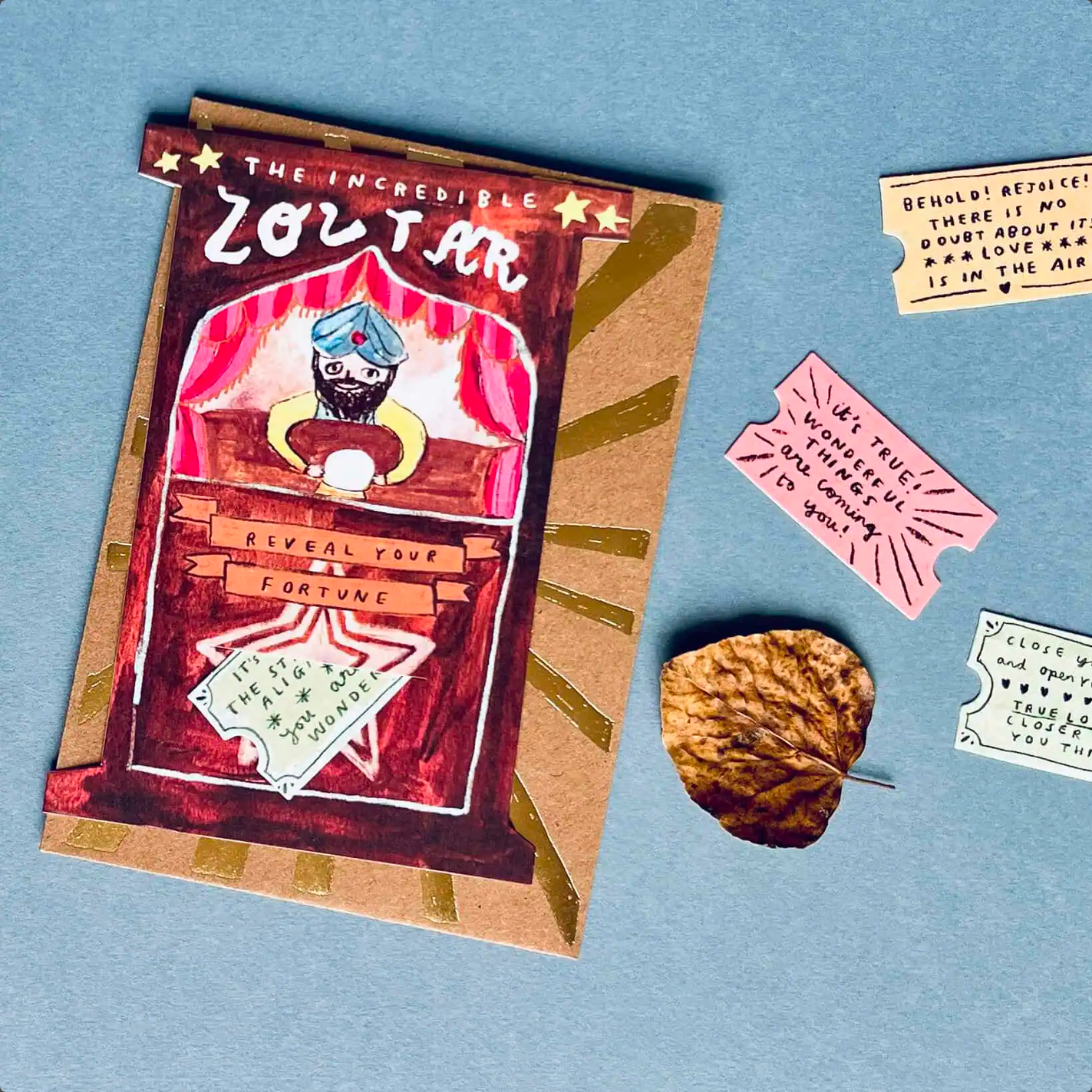 A hand-drawn zodiac card set displayed on a blue background, accompanied by a small dried leaf and sample ticket cards with whimsical text.