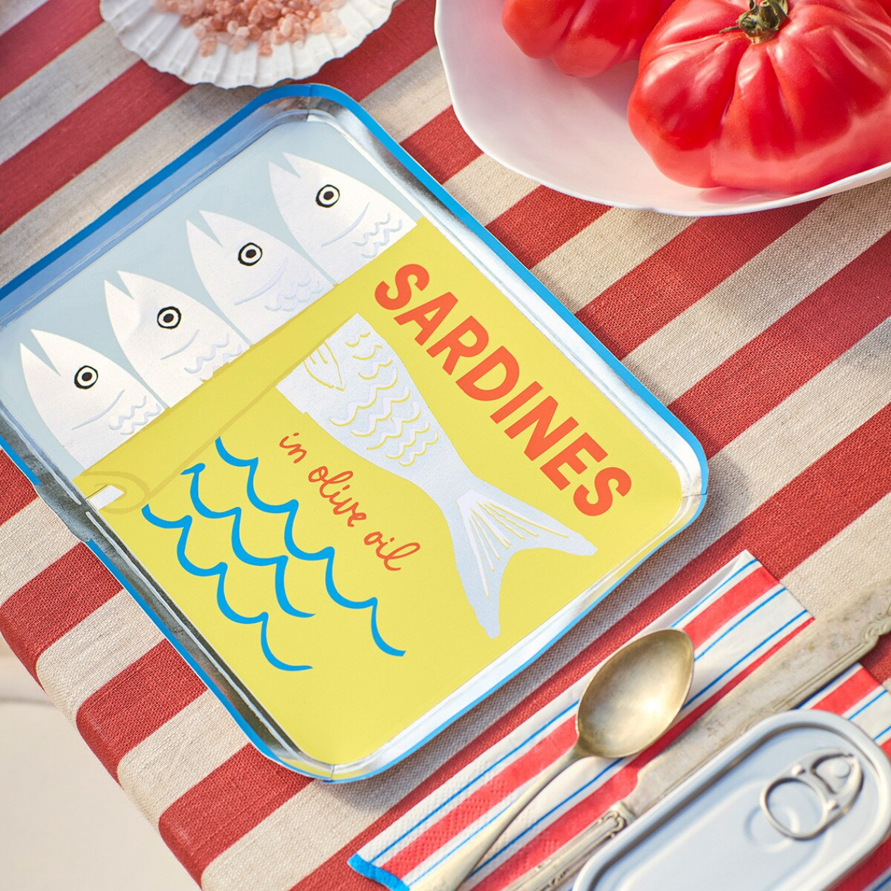 Sardine can-shaped phone case on a striped tablecloth with tomatoes and a spoon.