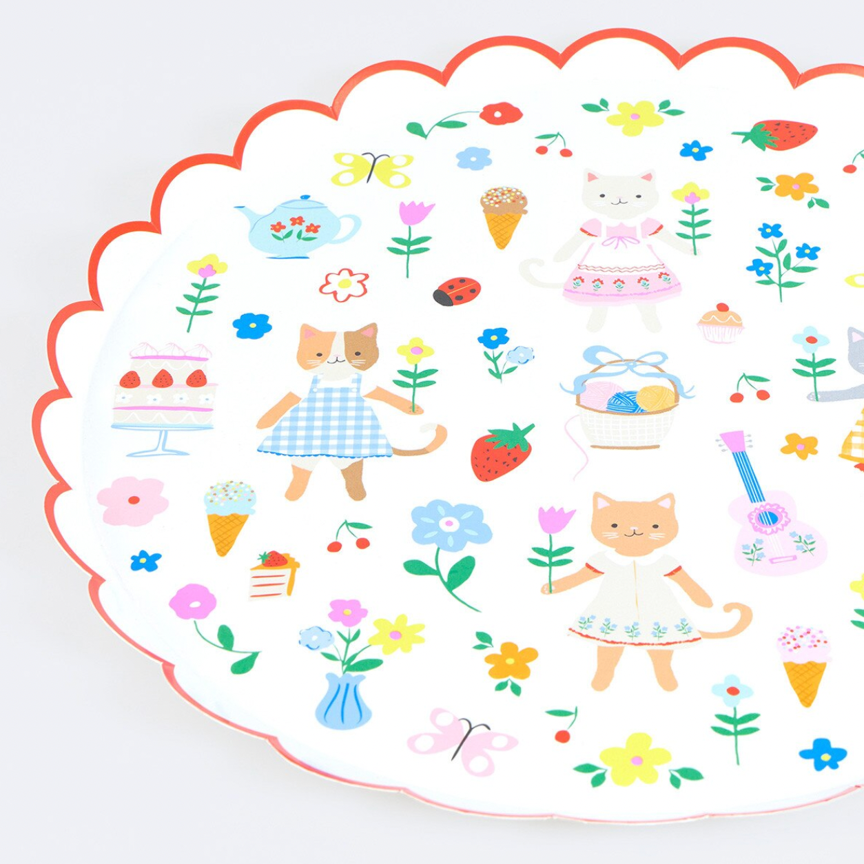 A scalloped white plate with a colorful whimsical print featuring two cats in dresses, flowers, strawberries, cakes, butterflies, and small illustrated objects, shown on a light neutral background.