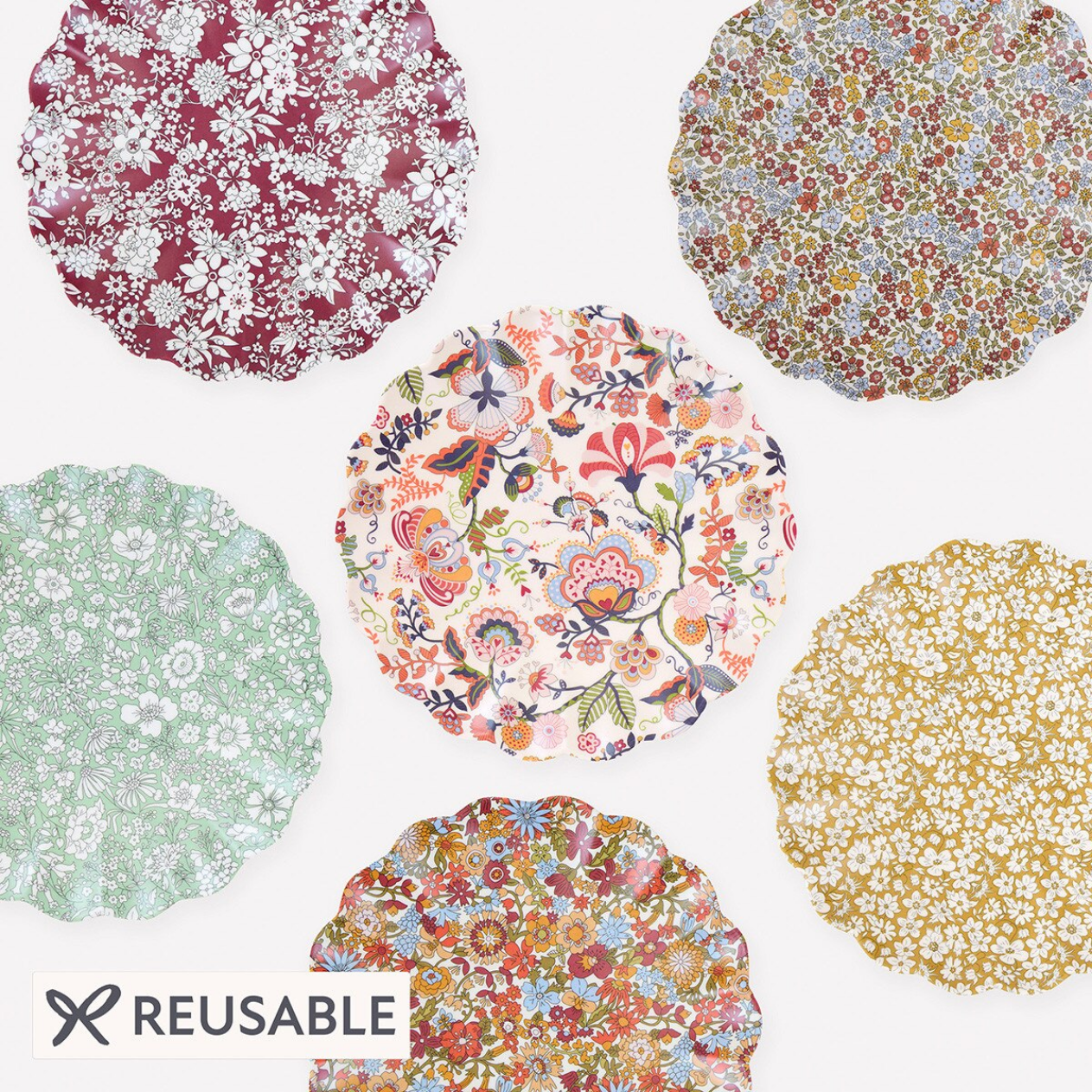 Six reusable plates with scalloped edges arranged on a white background, each featuring a different floral and botanical pattern in various colors. A 'REUSABLE' label with a scissors icon is in the bottom-left corner.