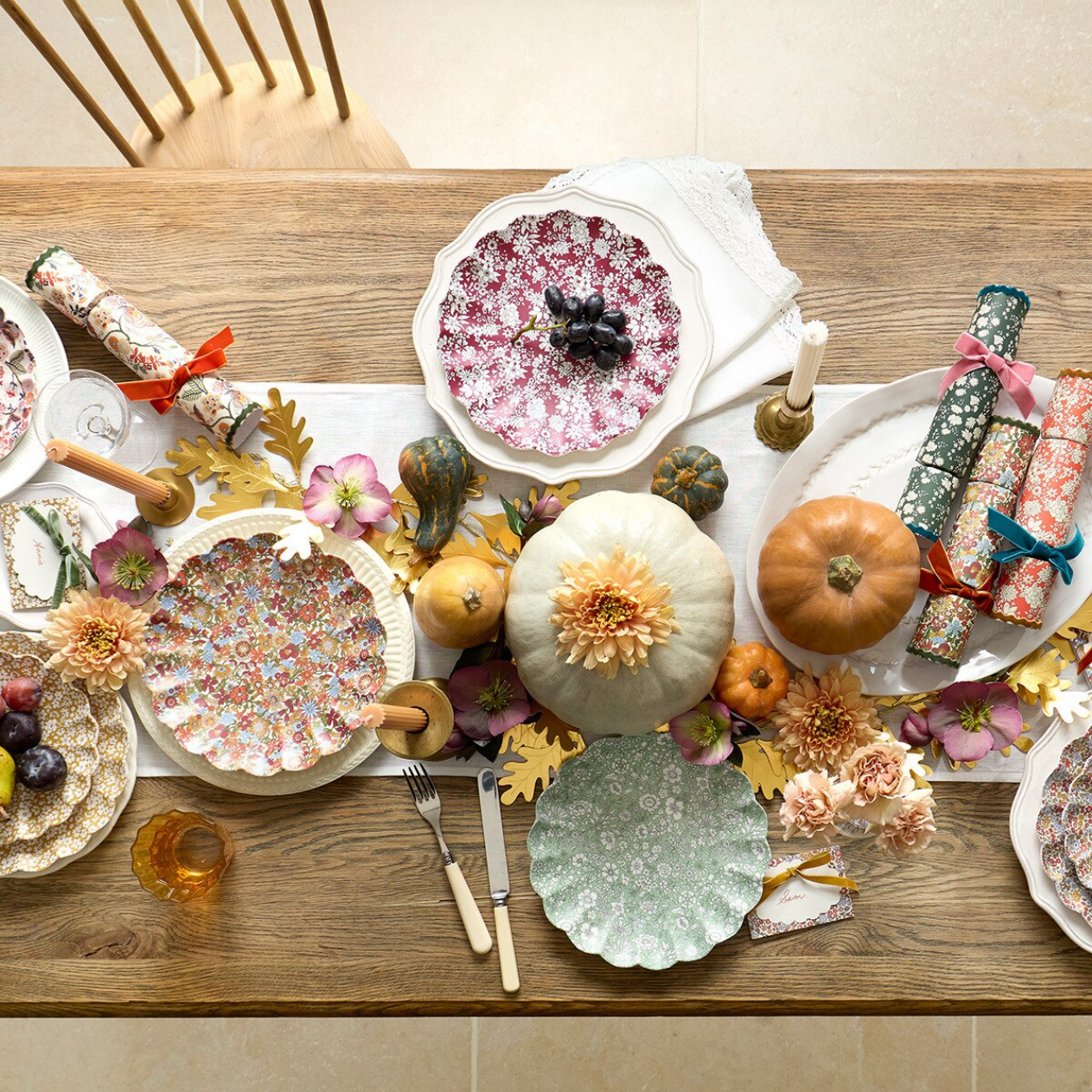 A styled autumn table setting with multiple decorative plates, patterned napkins, wrapped crackers, pumpkins, flowers, and a white runner on a wooden dining table.