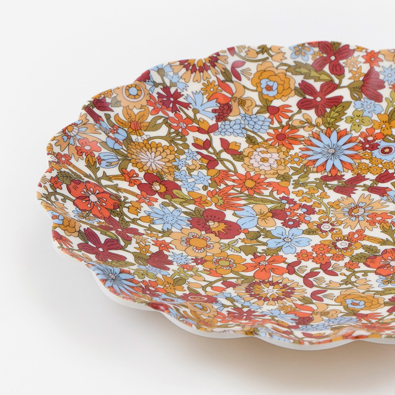 A close-up of a scalloped ceramic plate with a dense, multicolor floral pattern on a white background, photographed on a clean white surface.
