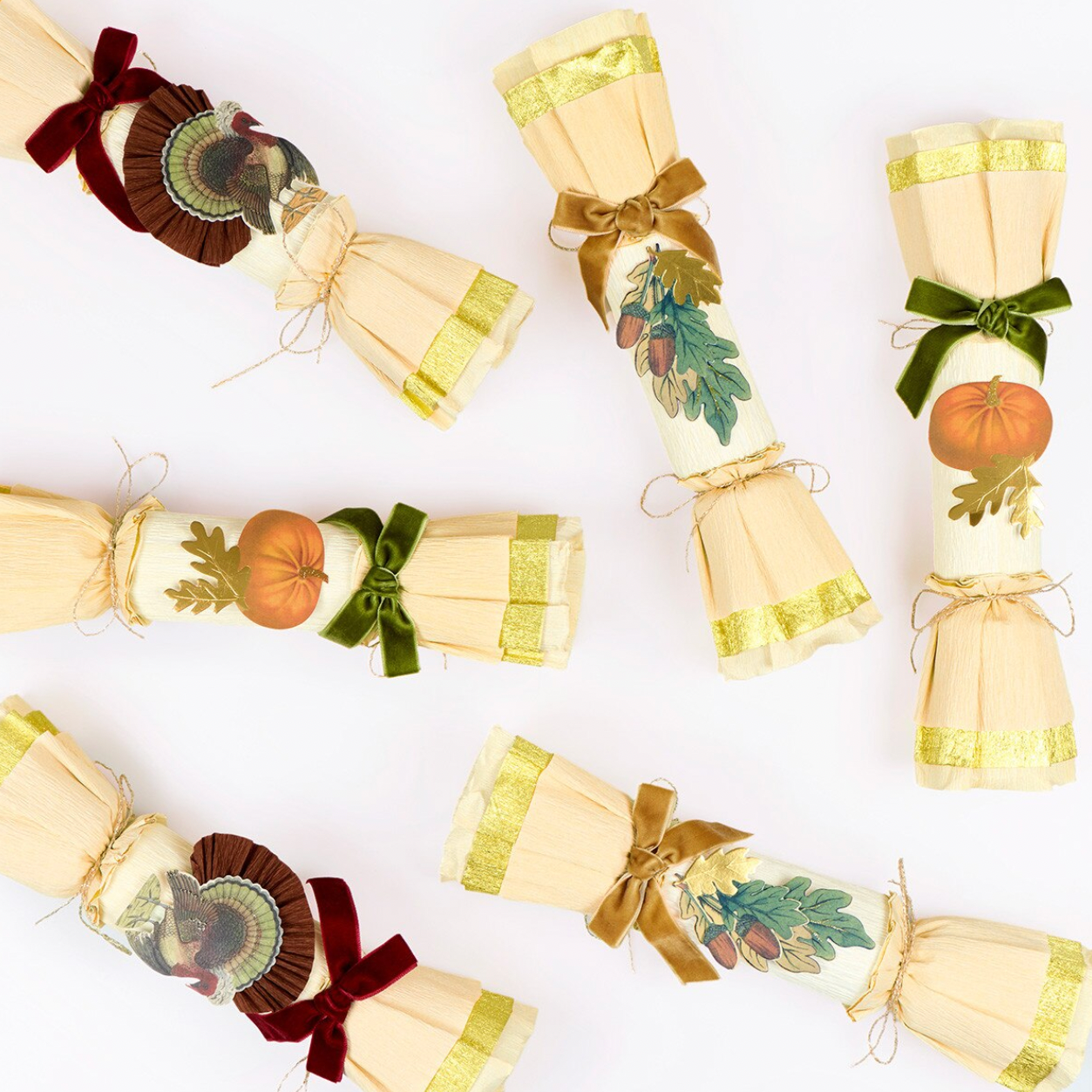 Five decorative paper cracker ornaments arranged on a white background, each with a fall harvest design including pumpkins, acorns, leaves, and a turkey motif, tied with ribbons in brown, olive green, and burgundy.