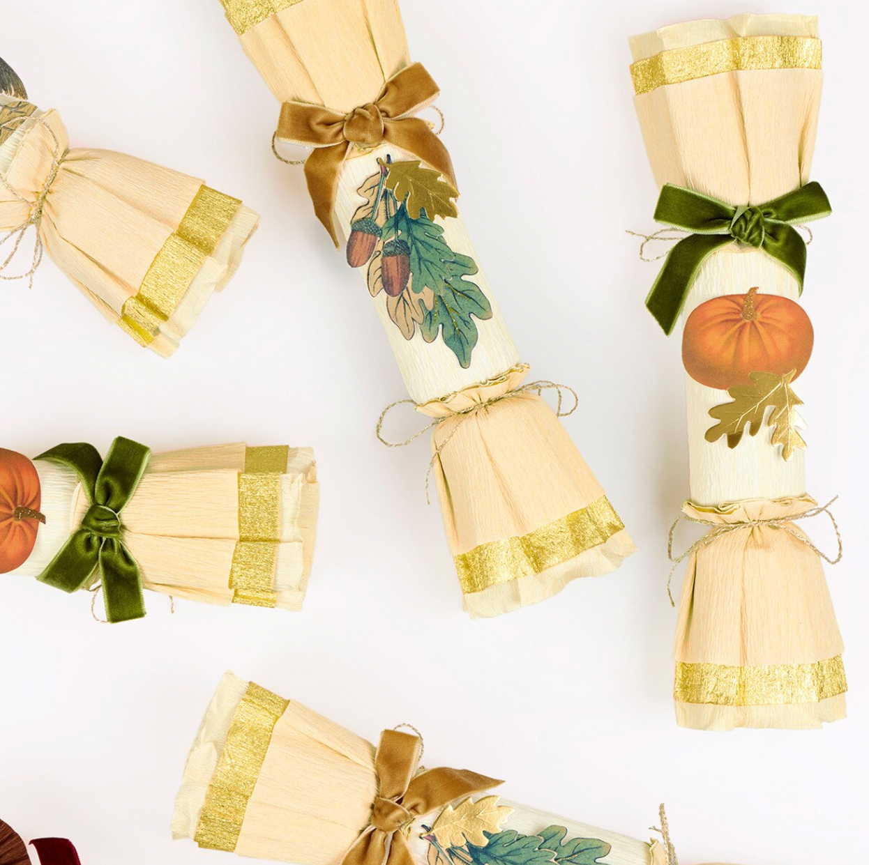 Three decorative cracker-style gift tubes arranged on a white background, each wrapped in cream paper with gold trim, ribbon bows, and autumn-themed printed illustrations including a pumpkin and green leaves.