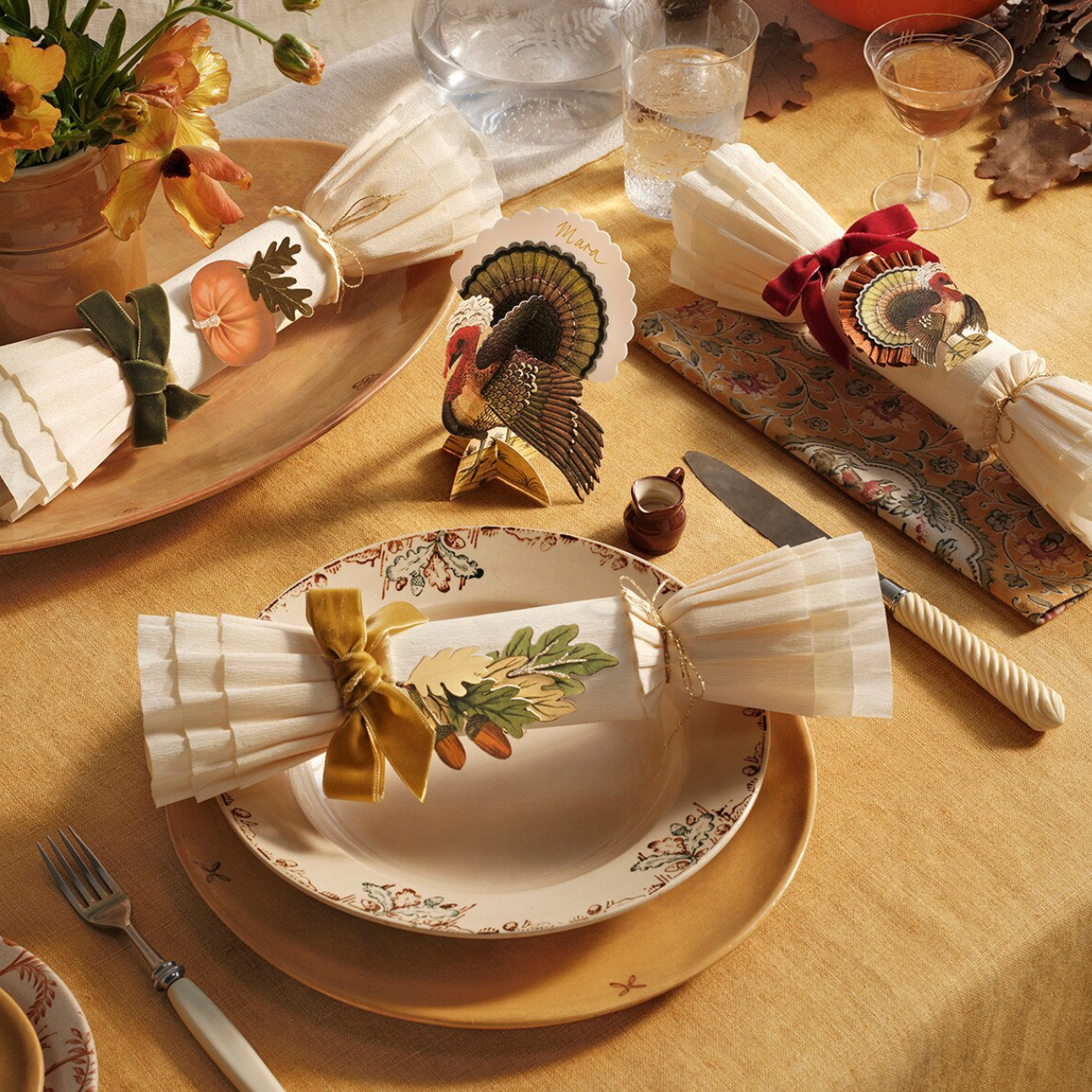 A styled Thanksgiving table setting with cream and gold patterned plates, folded white napkins tied with ribbon and bow, a decorative turkey figurine, a glass of water, and autumn table decor on a yellow tablecloth.