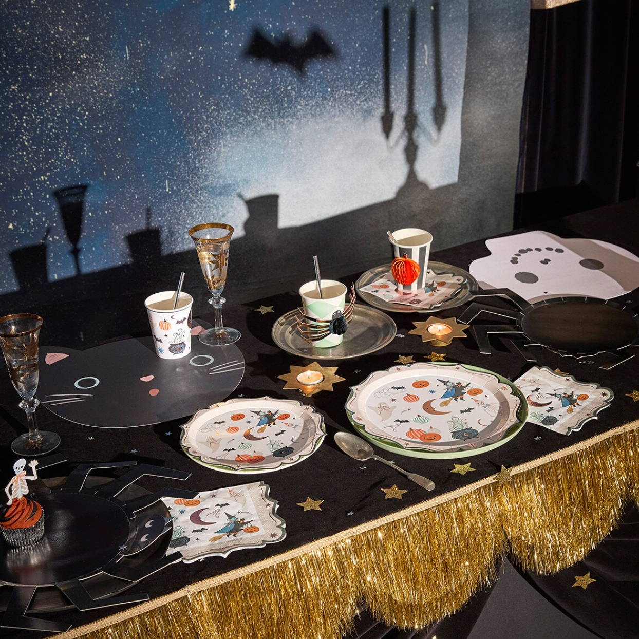 A Halloween-themed party table setting with black plates, cups, napkins, and glassware decorated with spider, cat, and moon motifs, arranged on a black tablecloth with gold fringe. A dark blue starry backdrop with bat and chandelier shadows is behind the table.