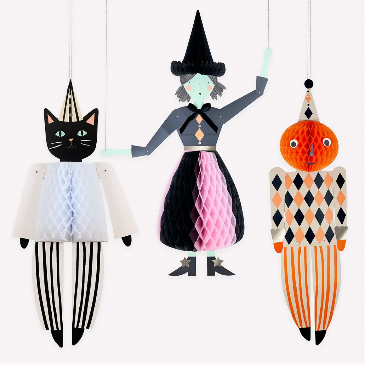 Three stylized hanging paper cutout figures on a plain white background: a black cat in a pointed hat and striped outfit, a witch figure in a black hat and pink-and-black dress, and an orange jack-o'-lantern-like figure in an orange, black, and cream harlequin pattern.