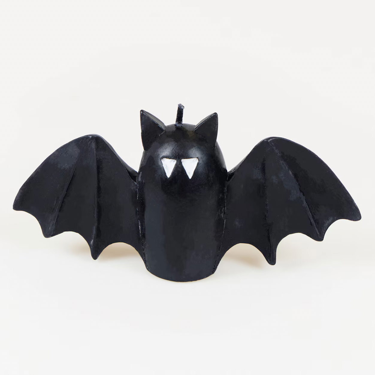 A black bat-shaped candle with wings spread wide, featuring pointed ears, a small wick at the top, and two white triangular eye shapes on the front, shown against a plain light background.