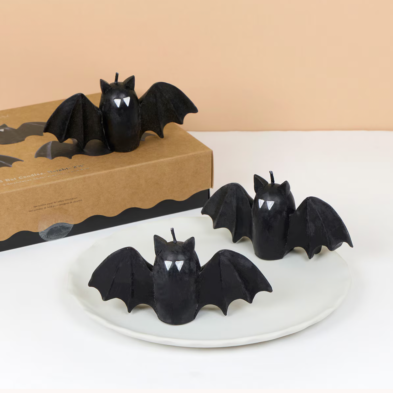 Three black bat-shaped candles with white eyes and teeth are displayed on a white plate, with a brown cardboard box in the background against a beige wall.