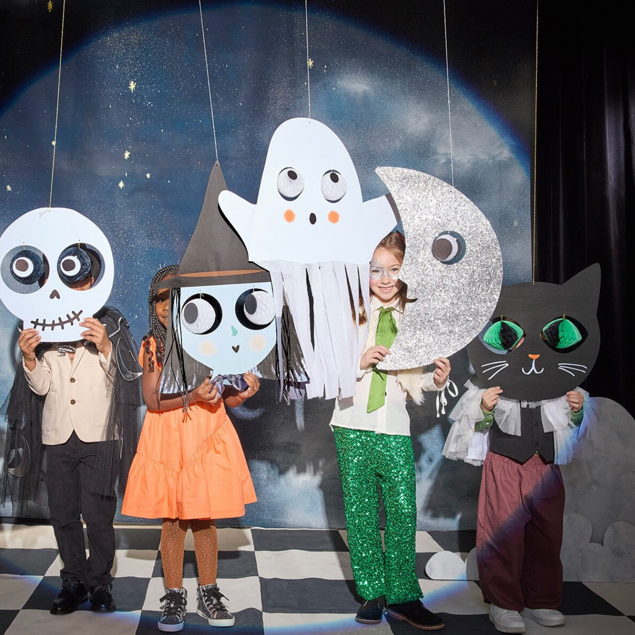 Four children wearing Halloween costumes stand in front of a moon and stars backdrop holding large paper cutout faces: a skeleton, a ghost, a crescent moon, and a black cat.