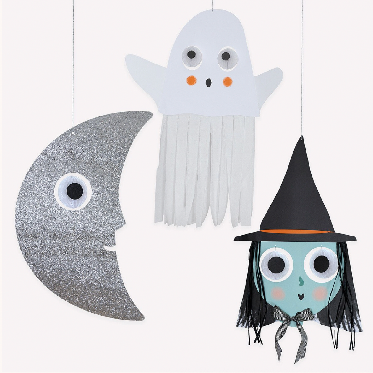 Three handmade Halloween hanging decorations made from cardstock and glitter: a silver glitter crescent moon with a googly eye, a white ghost with white fringe, and a teal witch with a black hat and black fringe.