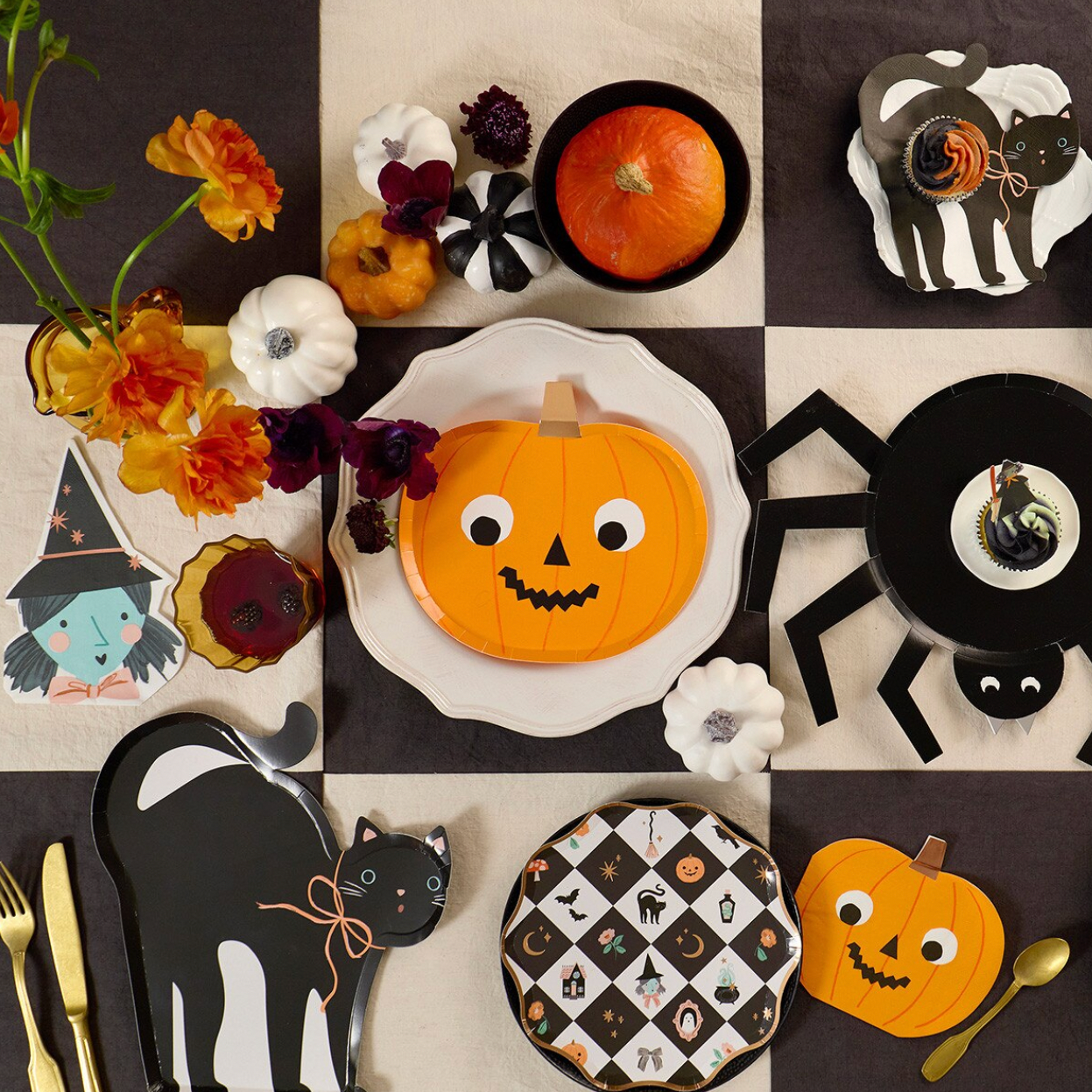 A styled Halloween tabletop flat-lay featuring decorative plates, cutlery, pumpkins, flowers, and black-and-white cat and spider cutout decorations arranged on a checkered dark brown and cream background.