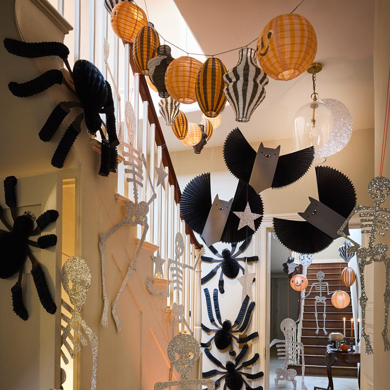 A Halloween-themed staircase decorated with black paper spiders, bats, and skeleton figures, orange and yellow patterned paper lanterns, and silver glitter skeleton decorations.