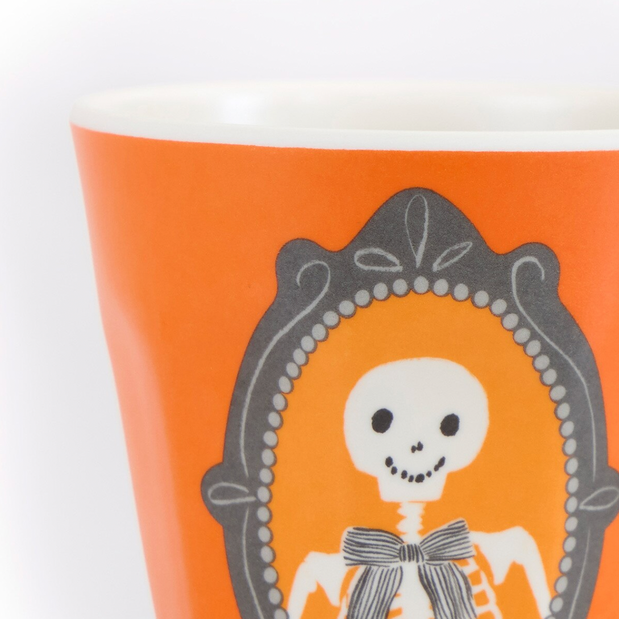 Close-up of an orange cup or tumbler with a white interior, featuring a decorative gray frame and a white skeleton illustration on the front.