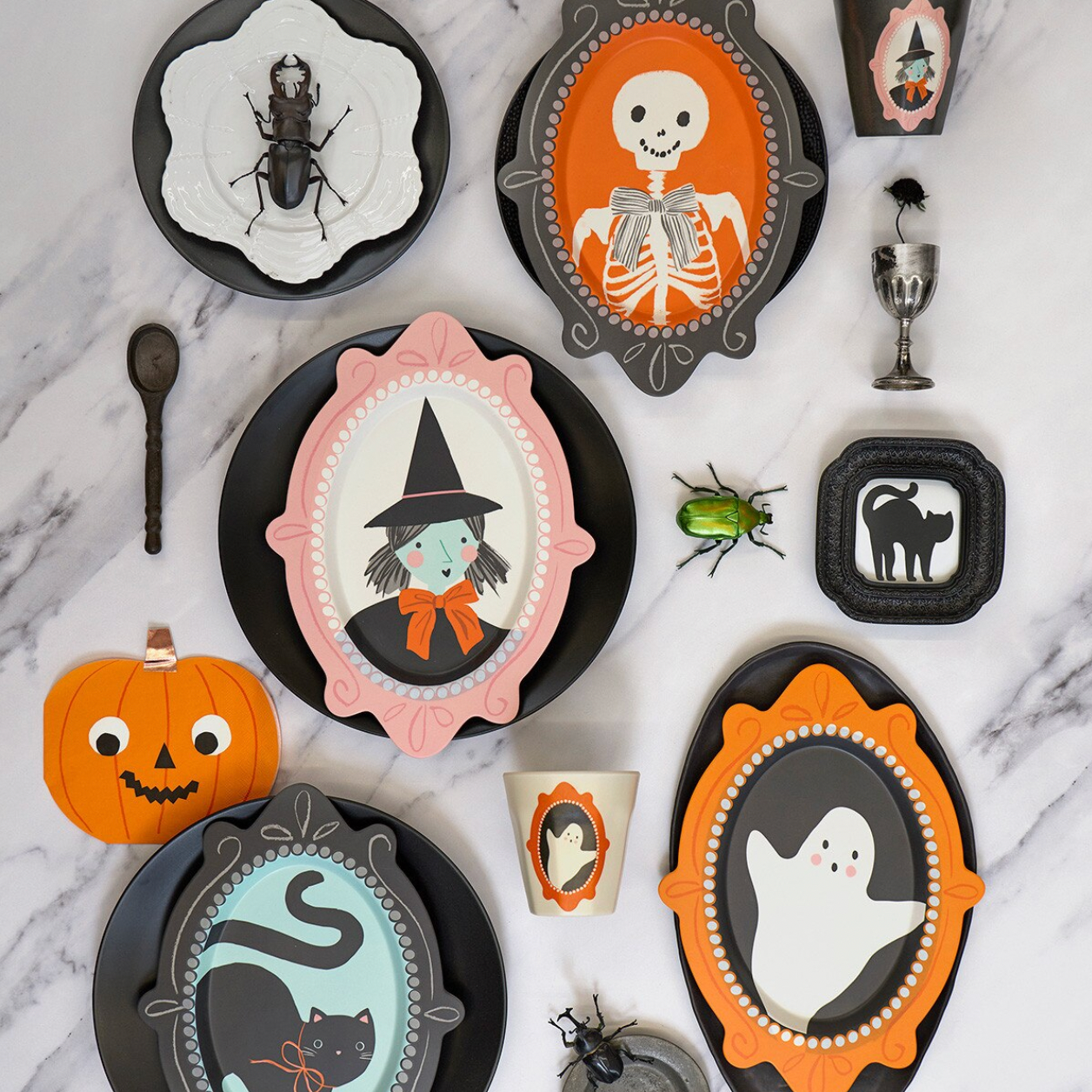 A styled flat-lay of Halloween-themed tableware and decor on a white marble surface, including black plates with orange, pink, and gray frames, a pumpkin, a beetle, a skeleton, a witch girl, a ghost, a black cat, a cup, and small decorative accessories.