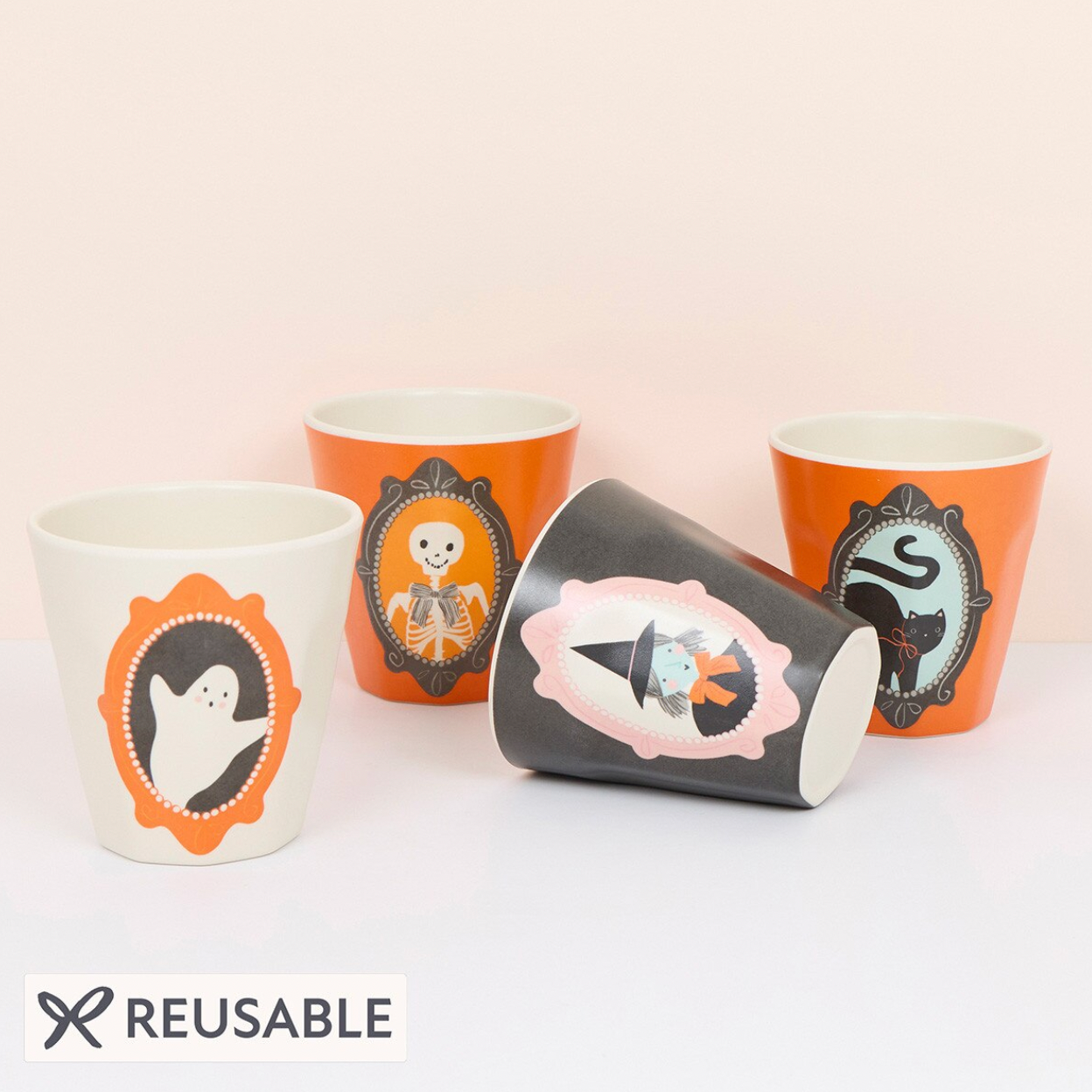 Four reusable Halloween-themed cups arranged on a white surface against a pale pink background. One white cup shows a black-and-orange ghost design, two orange cups show skeleton and cat designs, and one dark gray cup lies on its side showing a witch hat design.