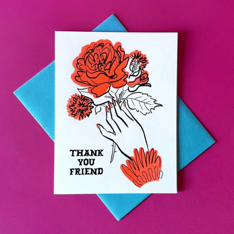 A square white greeting card with a black-and-red floral illustration and the words 'THANK YOU FRIEND' in black, placed on top of a blue envelope against a bright pink background.