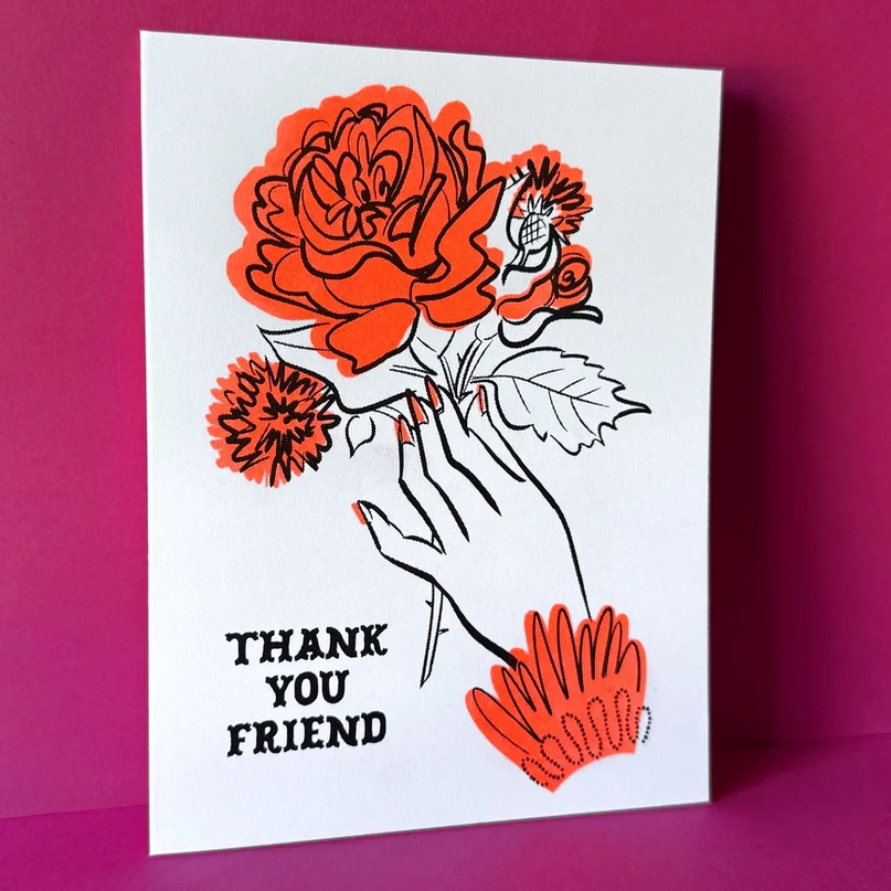 A white greeting card featuring a graphic illustration of a hand holding red-orange flowers, with the text 'THANK YOU FRIEND' in black. The card is displayed standing upright against a vibrant magenta background.