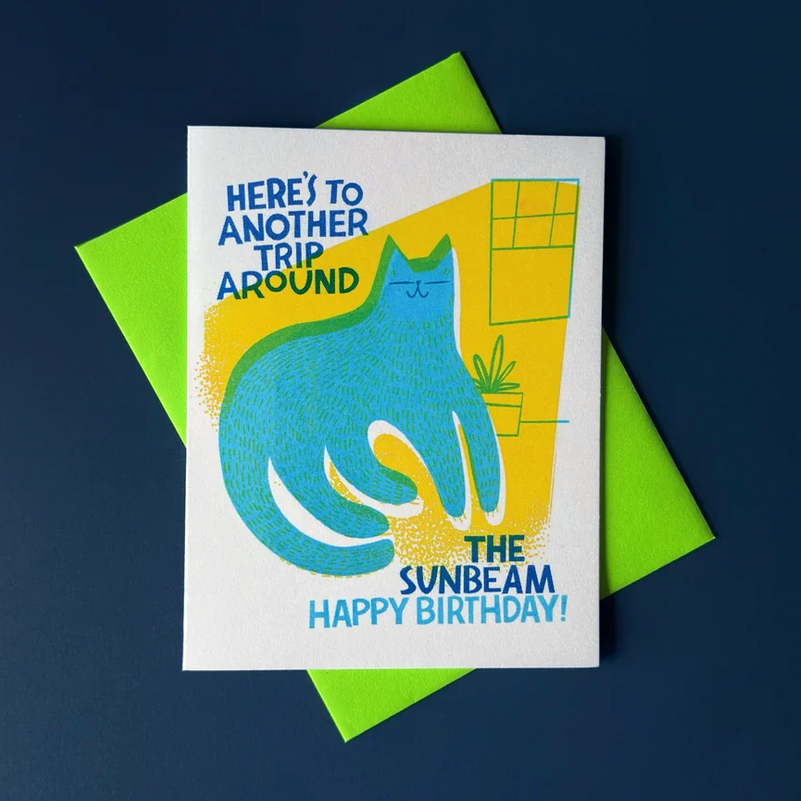 A square birthday card with a bright blue cat illustration and the text 'HERE'S TO ANOTHER TRIP AROUND THE SUNBEAM HAPPY BIRTHDAY!' is shown on top of a neon green envelope against a dark navy background.