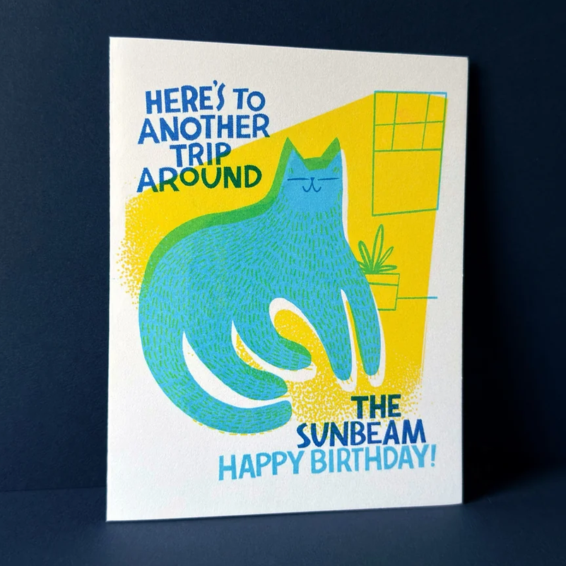 A square birthday card with a minimalist illustration of a blue cat under a yellow sunbeam, with the text 'HERE'S TO ANOTHER TRIP AROUND THE SUNBEAM HAPPY BIRTHDAY!', photographed against a dark navy background.