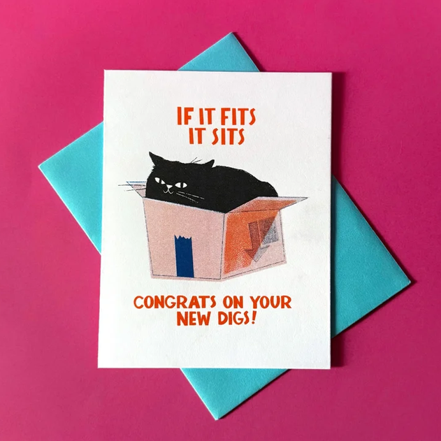 A white greeting card with a humorous illustration of a black cat sitting in a box, with orange text reading 'IF IT FITS IT SITS' and 'CONGRATS ON YOUR NEW DIGS!'. The card is displayed on top of a teal envelope against a bright pink background.